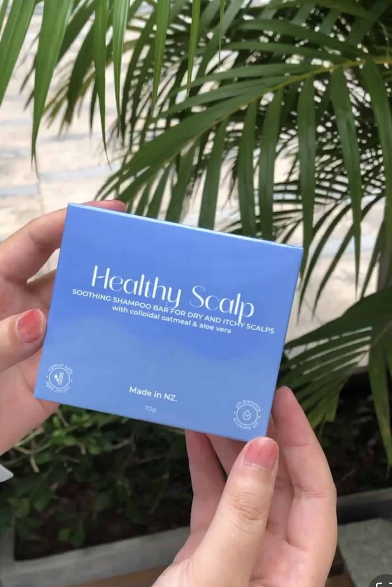 Hand holding a blue box labeled 'Healthy Scalp' with green foliage in the background